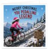 Female cyclist Christmas card showing a woman riding a road bike up a snowy mountain climb, wearing a bright pink cycling outfit and pink Santa hat. Snow-covered peaks, pine trees and falling snow create a festive winter scene. A wooden sign reads “EPIC VIEWS”, “WELL EARNED” and “CAKE AWAITS”. Large text across the top reads “MERRY CHRISTMAS, YOU PEDALLING LEGEND”.