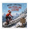 Male cycling Christmas card featuring a cheerful road cyclist climbing a snowy mountain on a red bike, wearing a festive red jersey and Santa hat over his helmet. Snow-covered peaks, pine trees and a winding road fill the winter scene. A wooden sign reads “Epic Views”, “Well Earned” and “Cake Awaits”. Bold red and green text reads “Merry Christmas, You Pedalling Legend”.
