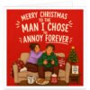 Funny Husband Christmas card with bold red background and cream text reading “Merry Christmas to the Man I Chose to Annoy Forever”. Festive illustration shows a couple in tartan pyjamas relaxing on a sofa, with the wife playfully poking her husband’s cheek as he holds a games controller. Includes a decorated Christmas tree, mugs, biscuits, candy cane, cosy blanket and “Love Laugh Annoy” sign.