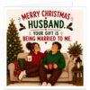 Funny Husband Christmas card featuring a cosy illustrated married couple relaxing on a sofa in matching red and green tartan pyjamas beside a decorated Christmas tree. The wife points playfully at her husband as they hold mugs. Front text reads “Merry Christmas Husband” and “Your Gift Is Being Married to Me”. Festive details include holly, baubles, fairy lights, greenery, berries, hearts, cushions and a candle lantern.