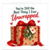 Funny romantic Christmas card for wife, girlfriend or partner featuring the text “You’re Still the Best Thing I Ever Unwrapped.” White background with dark green lettering and large red script, above a kraft-wrapped Christmas present decorated with red hearts, an oversized red ribbon and bow, mistletoe and a heart gift tag. Cheeky festive card from husband, boyfriend or partner.
