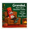 Funny Grandad Christmas card featuring the message "Grandad, You're Like a Christmas Cracker… Full of Bad Jokes and Strange Noises!" Colourful illustration of a smiling red Christmas cracker with grey moustache, black glasses, gold paper crown and green tartan slippers, relaxing in a brown armchair with a steaming mug. Cosy festive scene with decorated Christmas tree, wrapped presents, roaring fireplace and dark green background. Humorous Christmas greeting for Grandad, Grandpa or Granda.