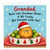Funny Grandad Christmas card featuring the message "Grandad, You're Like Christmas Dinner… A Bit Crusty, But Everyone Loves You!" in bold red and black lettering. Colourful illustration of a grumpy roast potato wearing a Santa hat, surrounded by Brussels sprouts and gravy in a festive roasting dish on a red checked tablecloth. Light blue background with white snowflakes. A cheeky, humorous Christmas card for Grandad, Grandpa or Grandfather from grandchildren.