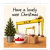 Northern Ireland Christmas card featuring the greeting "Have a lovely wee Christmas" in playful black lettering. The festive Belfast design showcases the iconic yellow Harland and Wolff shipyard cranes, Samson and Goliath, alongside a miniature decorated Christmas tree, a white mug featuring the red Belfast skyline and a wrapped present with holly. Set against a soft cream background with a wooden tabletop, this charming square Christmas card celebrates Northern Irish heritage and festive cheer.